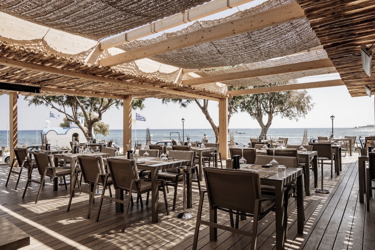 Shaded beachfront dining terrace with wooden tables, brown chairs, and woven pergola overlooking the Aegean Sea at NEMA Design Hotel & Spa.