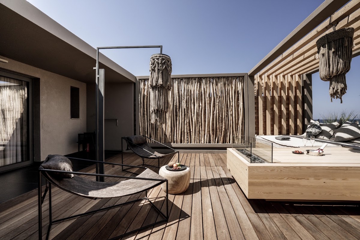 Modern beachfront terrace with wooden decking, black loungers, macramé wall panels, hanging decorations, and a sunken seating area under clear sky.