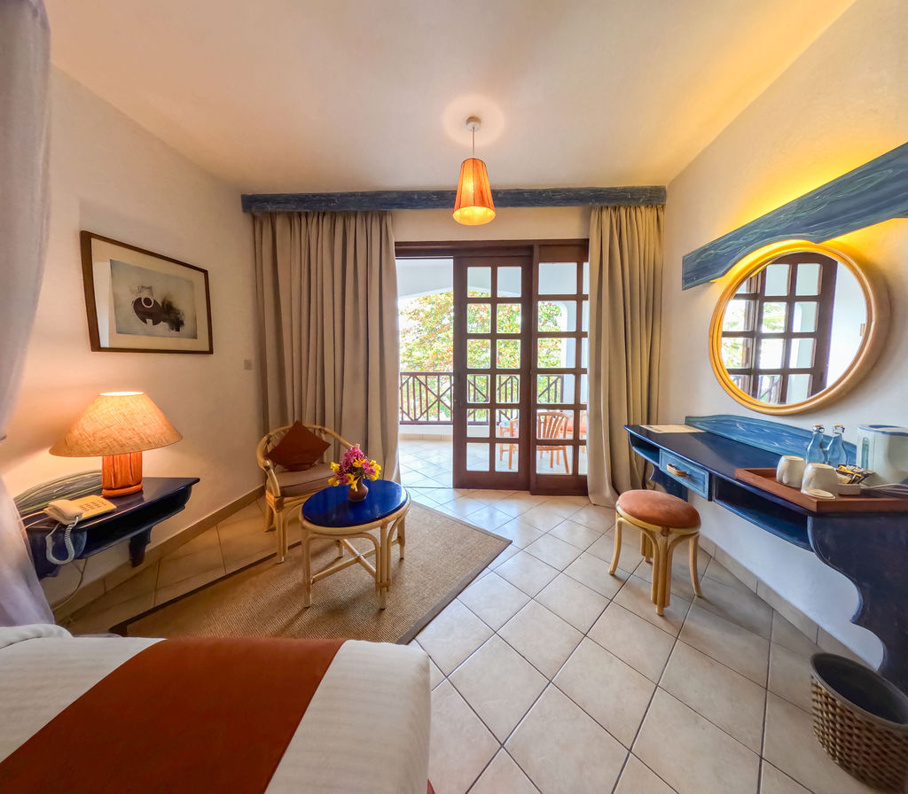 Resort room at Neptune Beach Resort with sitting area, blue round table, and French doors opening to a balcony overlooking greenery.