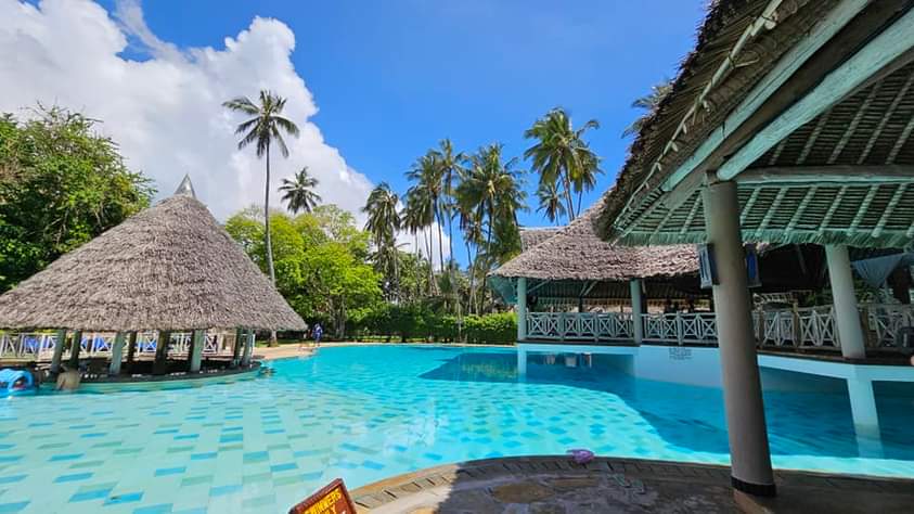 Large turquoise pool with thatched-roof structures, palm trees, and covered pavilions at Neptune Beach Resort under blue sky.