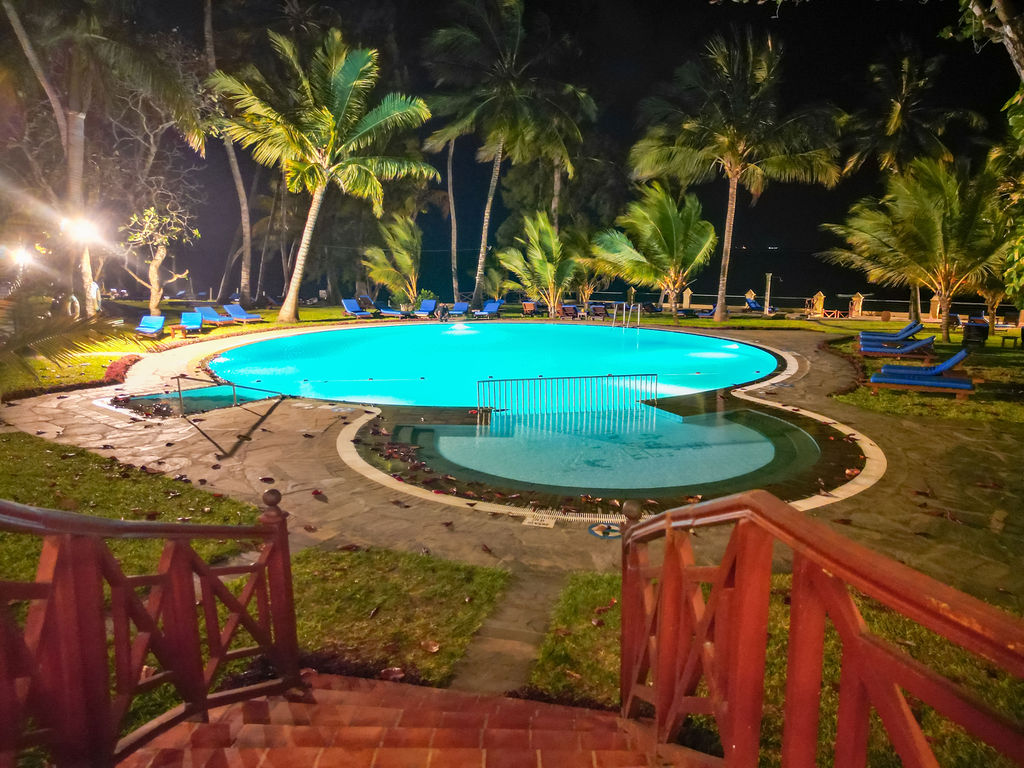 Illuminated resort pool with turquoise water surrounded by palm trees, lounge chairs, and manicured lawn at night.