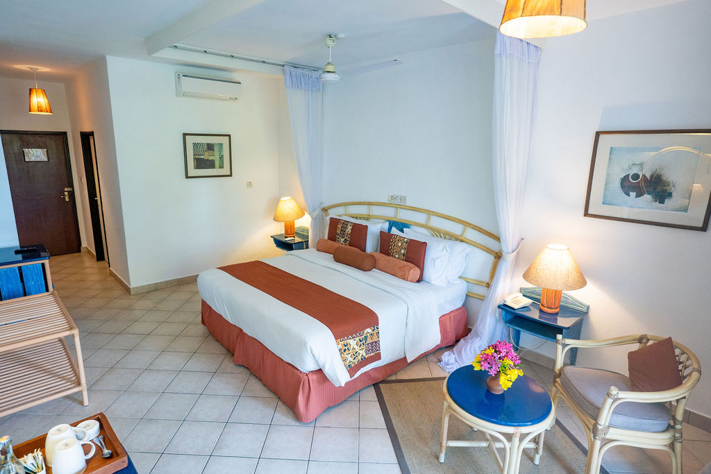 Spacious resort bedroom with queen bed, coral bedding, sheer curtains, two bedside lamps, tile flooring, and seating area with blue side table holding pink flowers.
