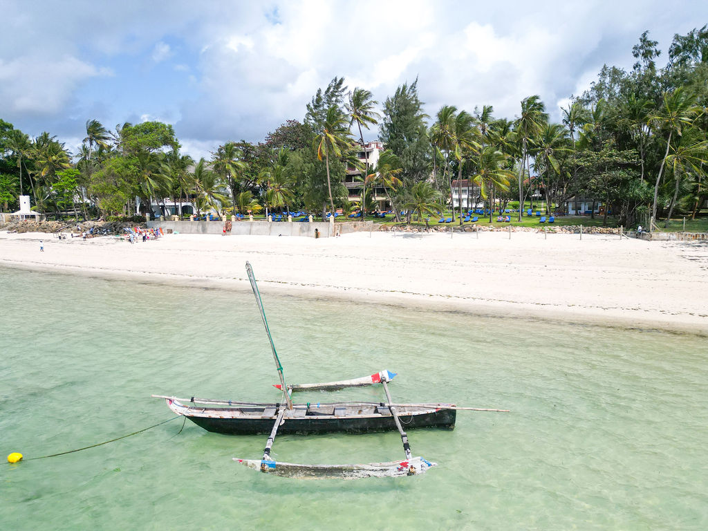 Outrigger boat in turquoise water off Neptune Beach Resort's sandy shore lined with palm trees and resort buildings.