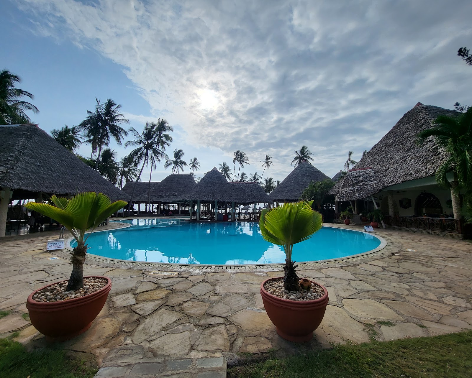 Illuminated thatched-roof pavilions with columns overlooking a large pool at dusk, with palm trees and beach umbrellas visible in the background.