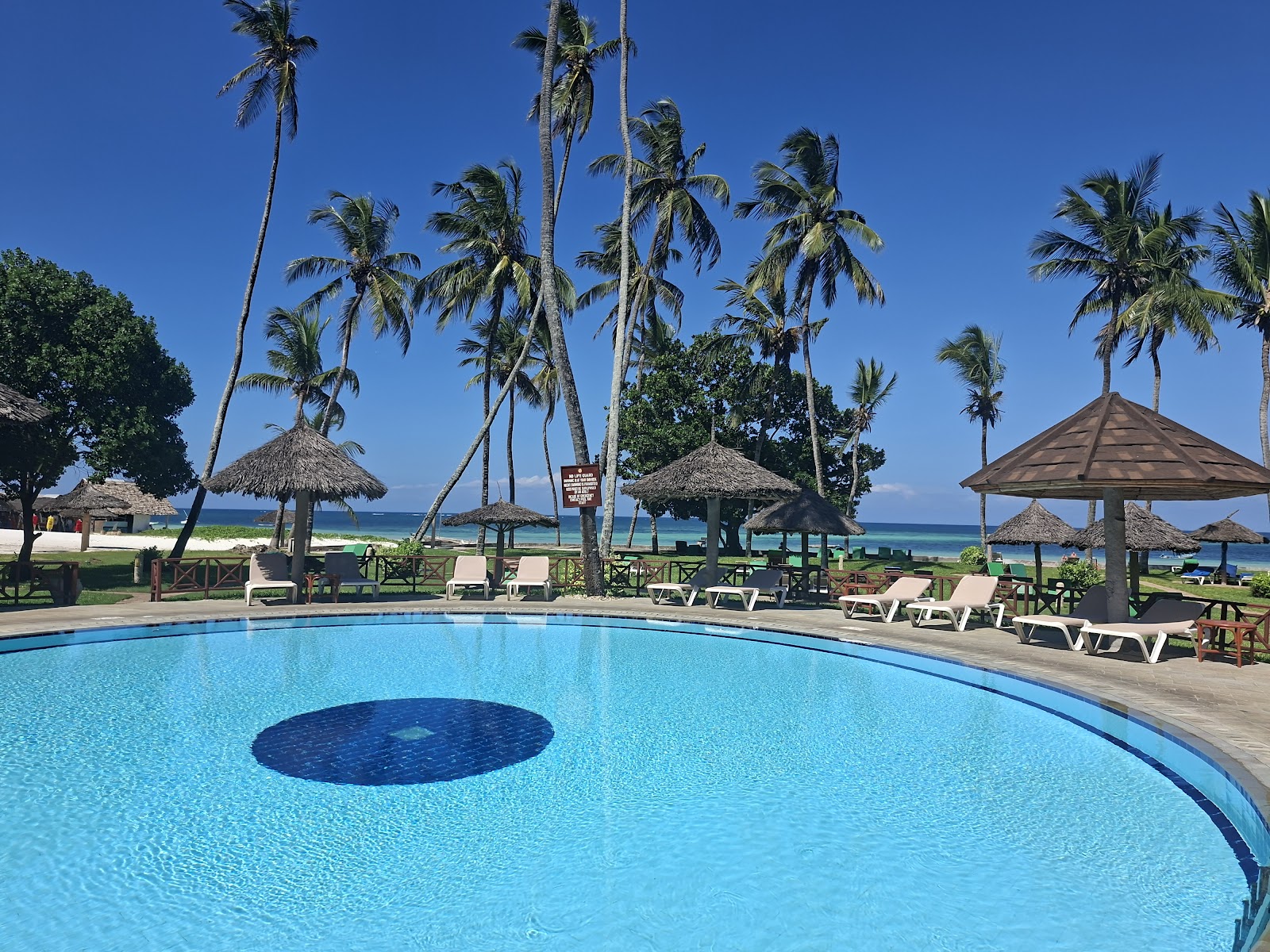 Illuminated thatched-roof pavilions with wooden columns line a turquoise pool at dusk, palm trees visible behind at Neptune Paradise Beach Resort & Spa.