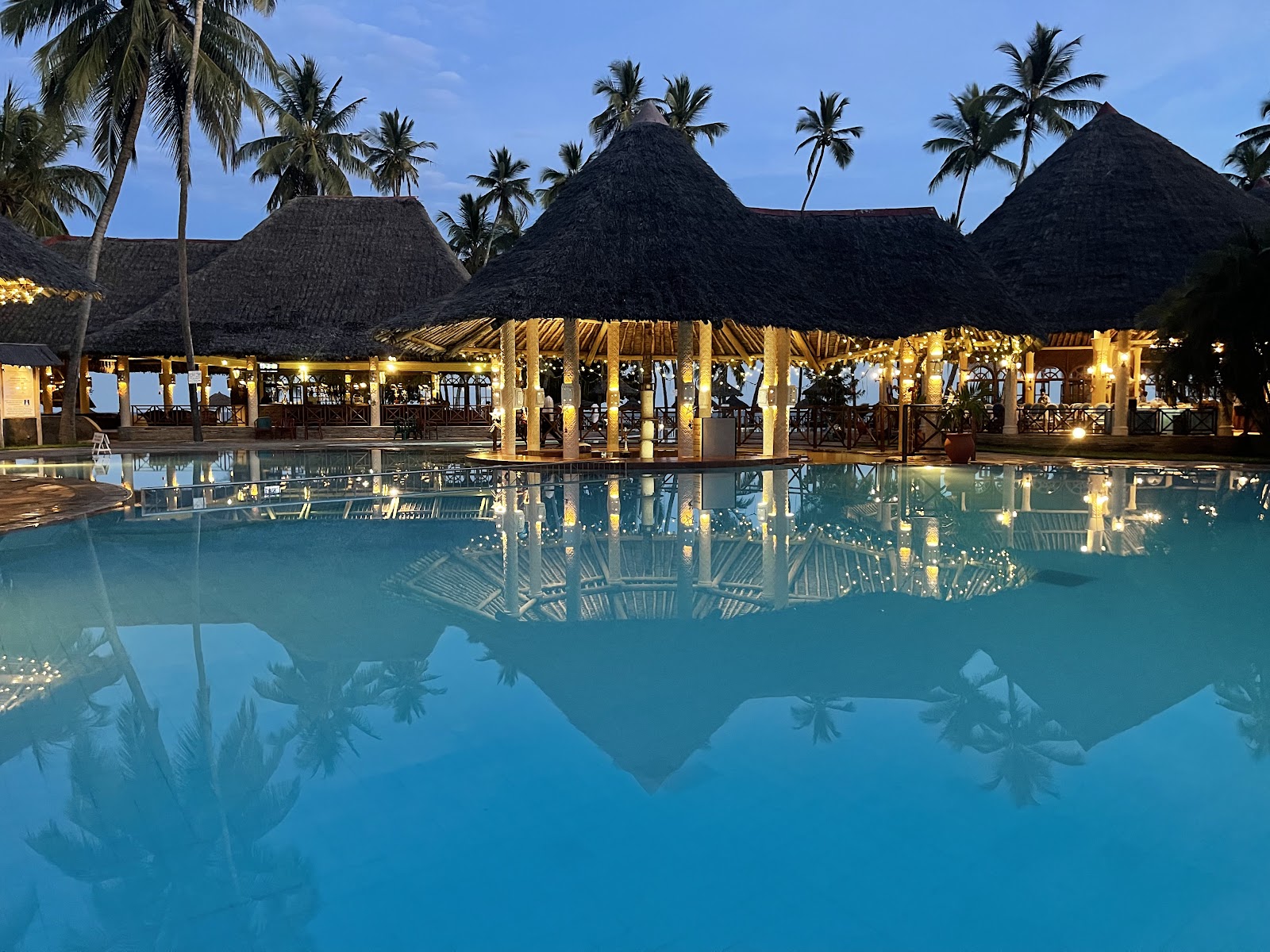 Blue swimming pool with thatched-roof bungalows, palm trees, and guests on loungers at Neptune Paradise Beach Resort & Spa.
