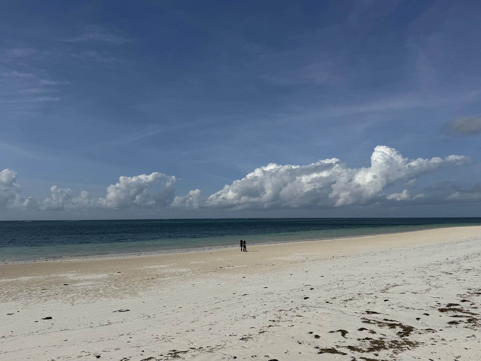 Two people stand in the shallow water off a sandy beach at Neptune Paradise Beach Resort & Spa under a blue sky with white clouds.