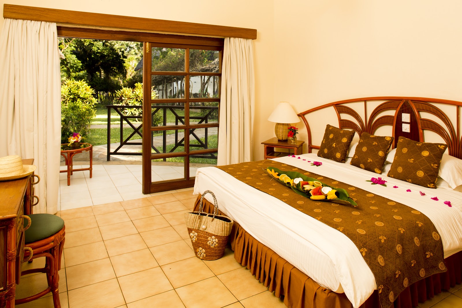 Resort bedroom with wooden bed frame, patterned bedspread, and flower petals, opening to terrace with lawn and trees beyond.