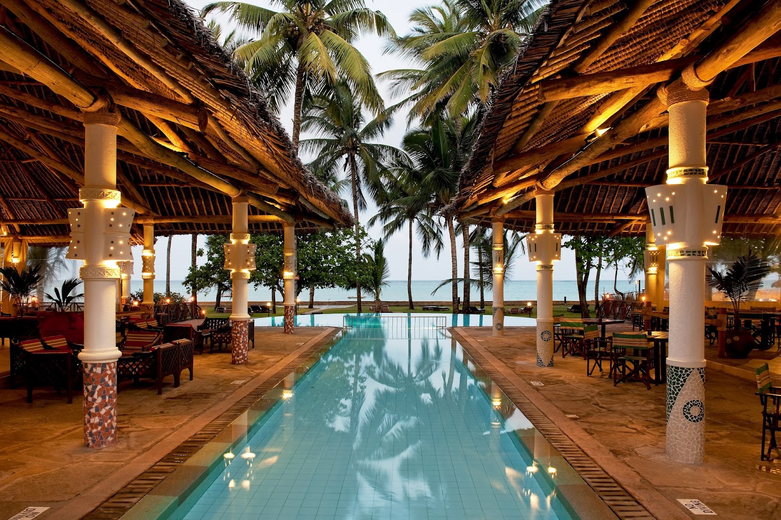 Long lap pool beneath a wooden pergola with white columns at Neptune Village Beach Resort & Spa, palm trees and ocean visible beyond.