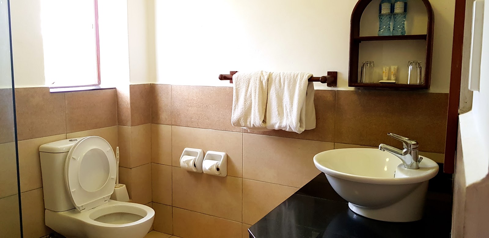 Bathroom with tan tile walls, white toilet, round sink, towel rack, soap dispensers, and wooden wall shelving at Neptune Village Beach Resort & Spa.