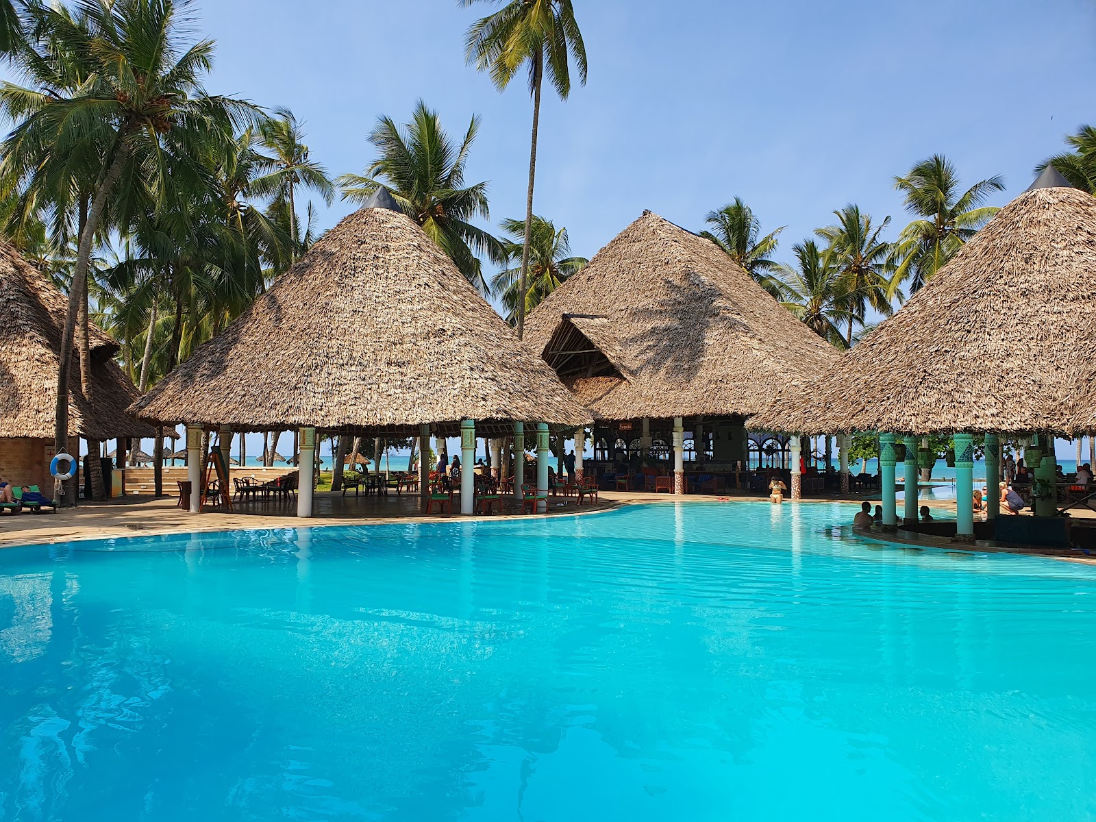 Large thatched-roof pavilion with wooden beams and white columns, furnished with red cushioned chairs and wooden benches, overlooking a pool and resort grounds.