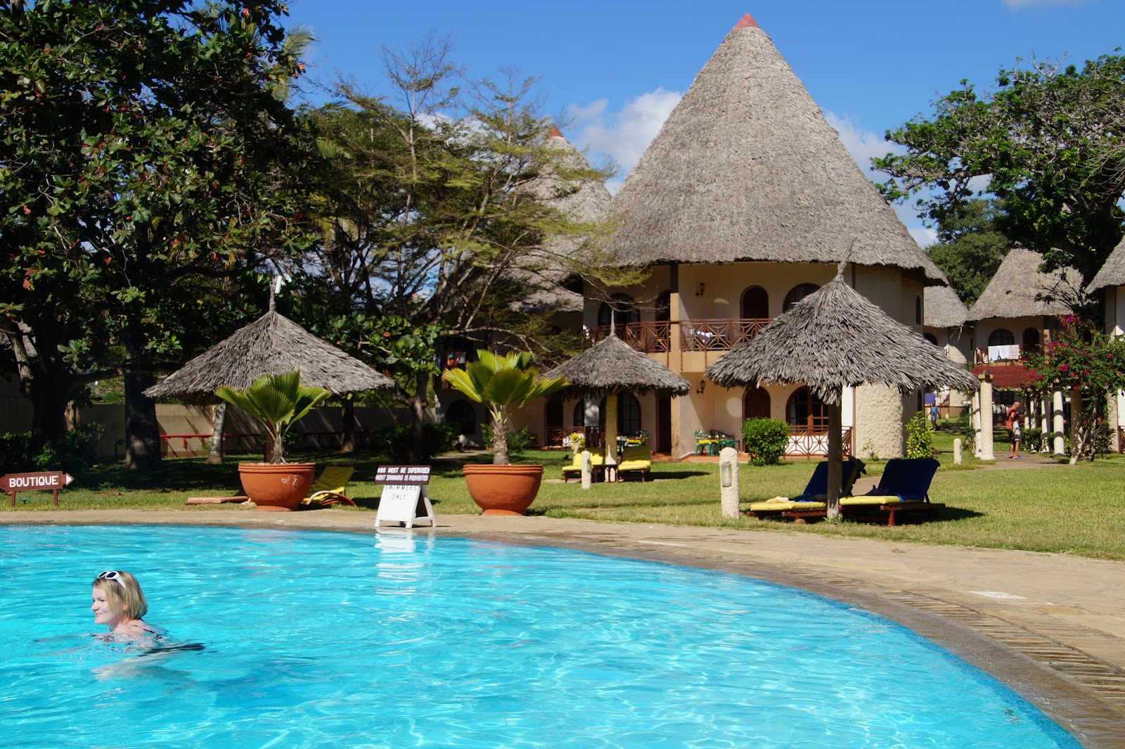 Turquoise pool with wooden deck surrounded by tall palm trees, manicured lawn, and ocean view at Neptune Village Beach Resort & Spa.