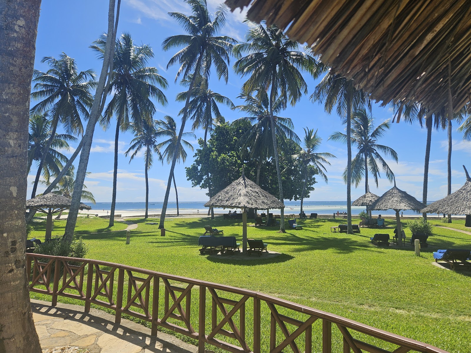 Traditional wooden dhow with white sail anchored in shallow turquoise water off a sandy beach lined with palm trees.