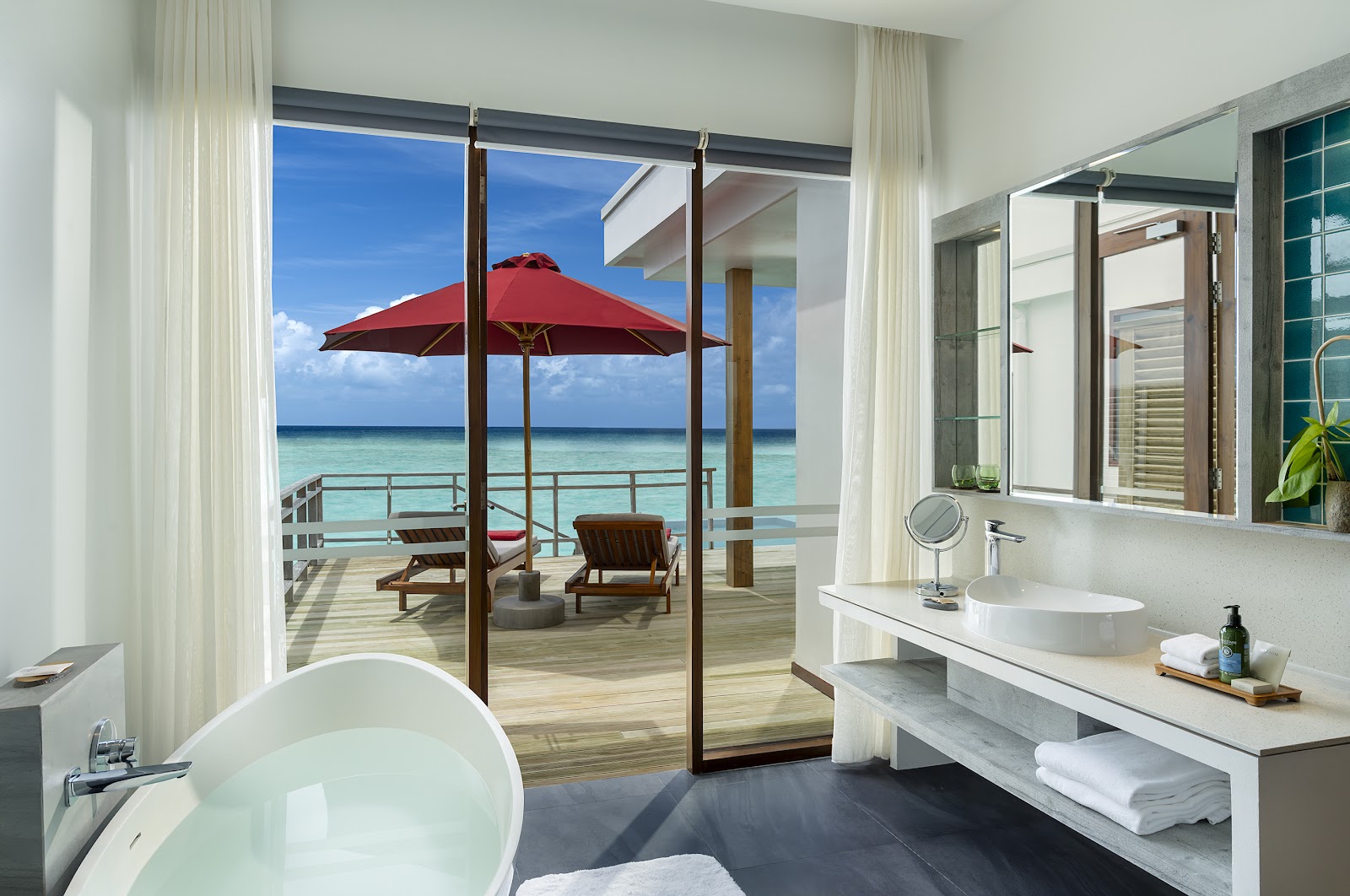 Bathroom with round soaking tub opens to a teak deck with red umbrella, lounge chairs, and turquoise ocean at NIVA Dhigali Maldives.