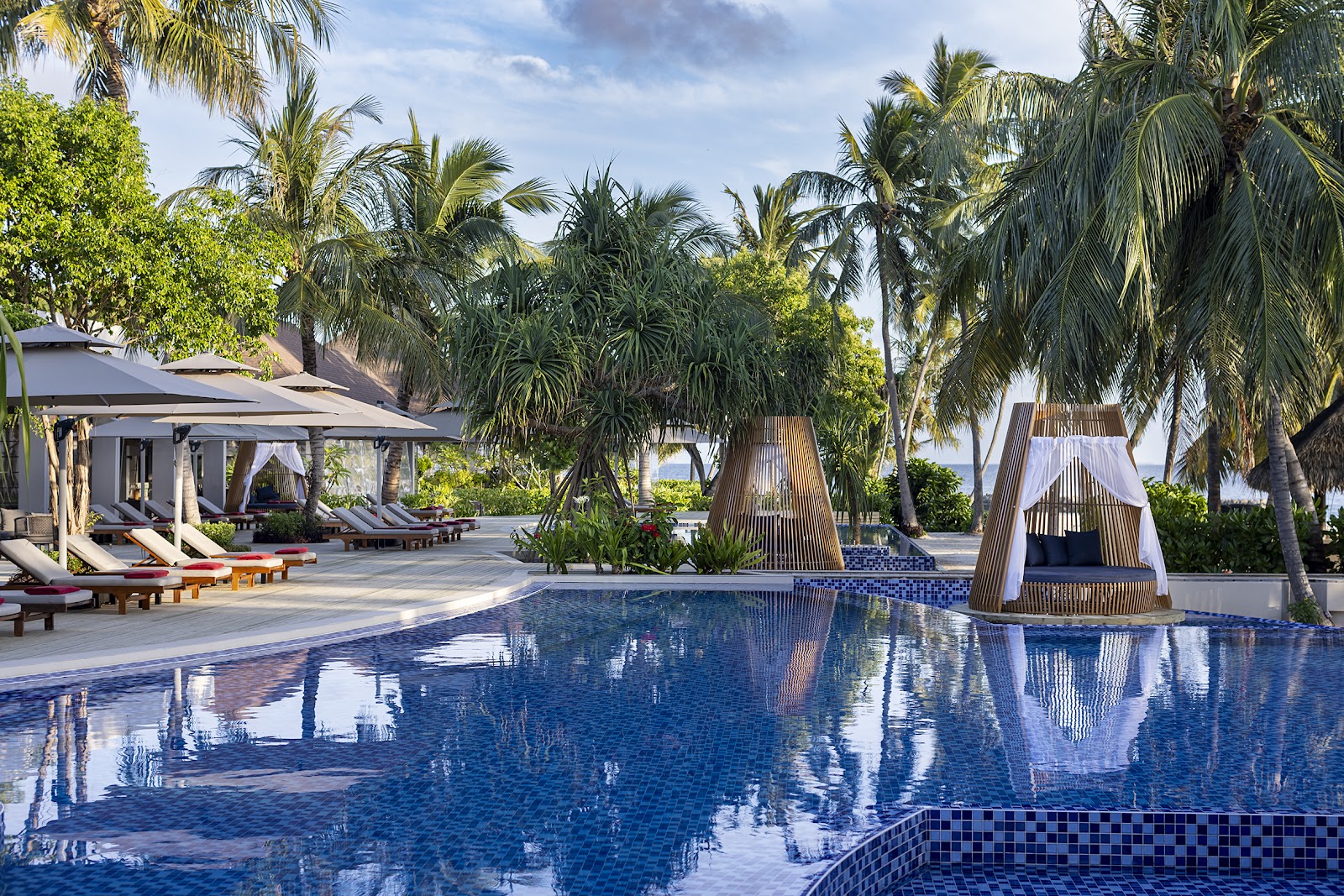 NIVA Dhigali Maldives pool with thatched pavilions, loungers under gray umbrellas, palm trees, and ocean view beyond.