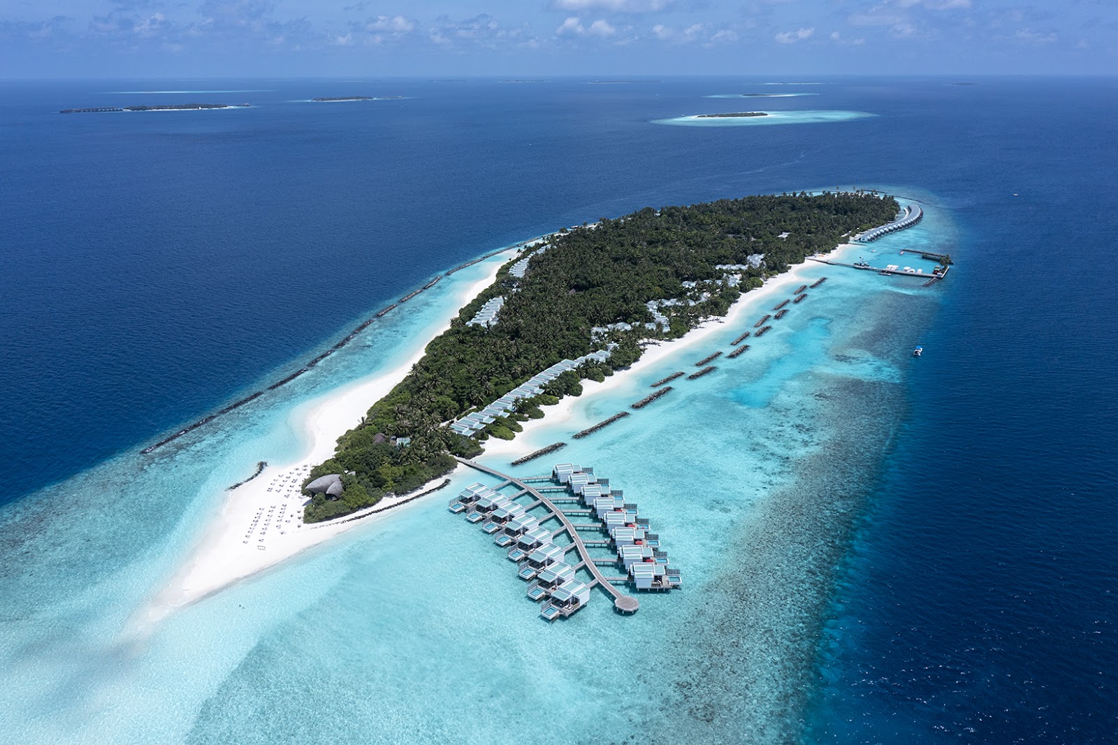 Aerial view of NIVA Dhigali Maldives with overwater bungalows arranged in rows, white sand beach, dense vegetation, and turquoise lagoon surrounded by deep blue ocean.