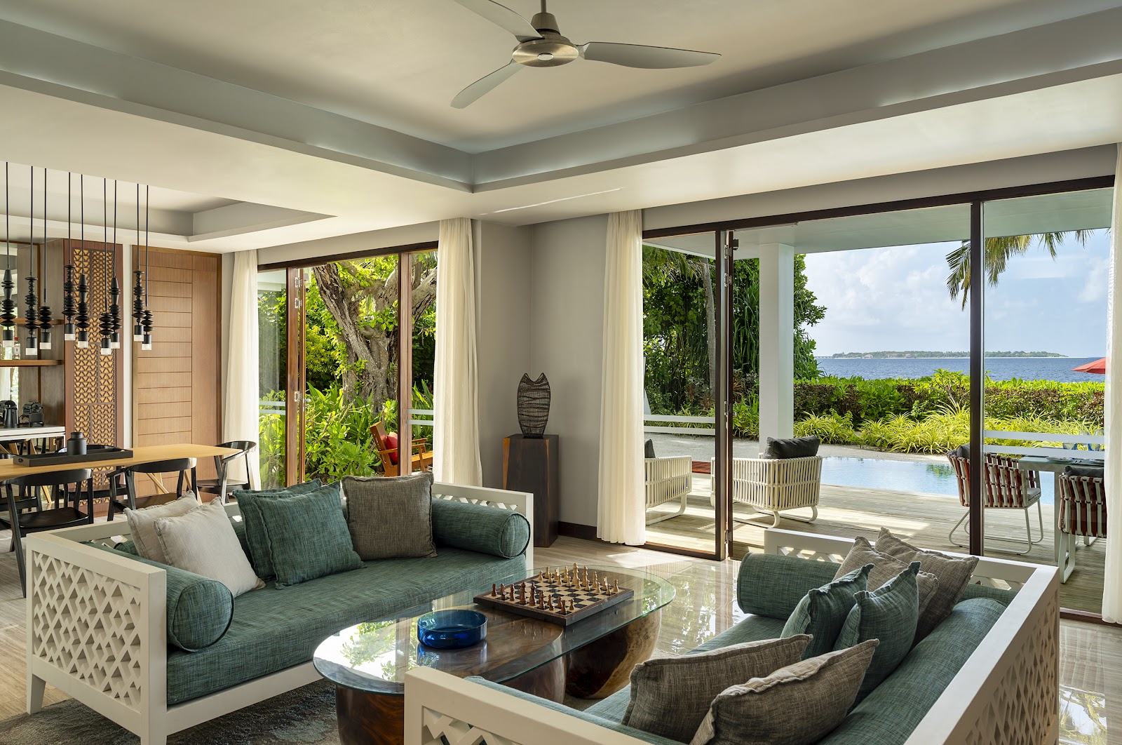 Modern resort villa interior with green upholstered seating, ocean view through floor-to-ceiling windows, and tropical landscape beyond.
