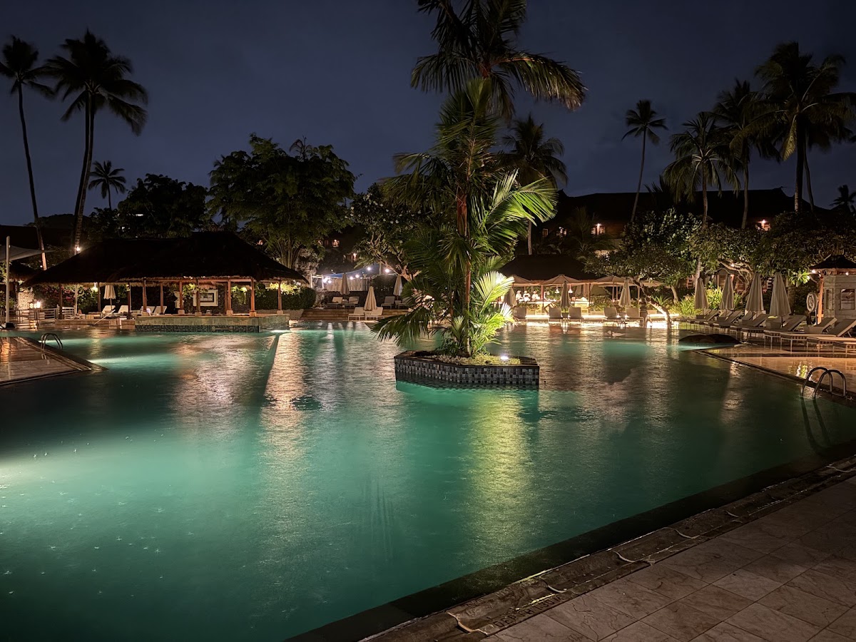 Illuminated turquoise pool at dusk with thatched-roof bungalows, palm trees, and lounge chairs surrounding the Nusa Dua Beach Hotel & Spa.