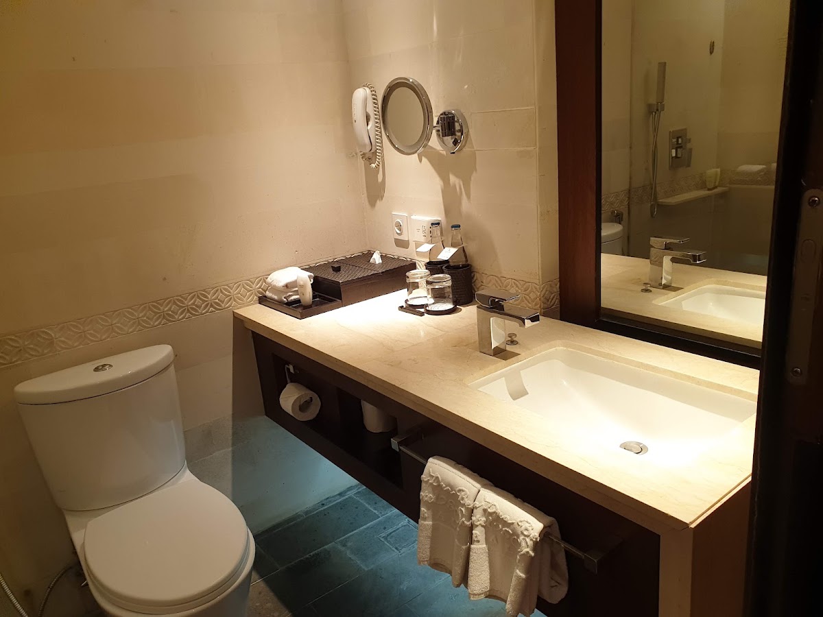 Modern resort bathroom with beige walls, dark wood vanity, white countertop sink, illuminated mirrors, and tiled floor at Nusa Dua Beach Hotel & Spa.