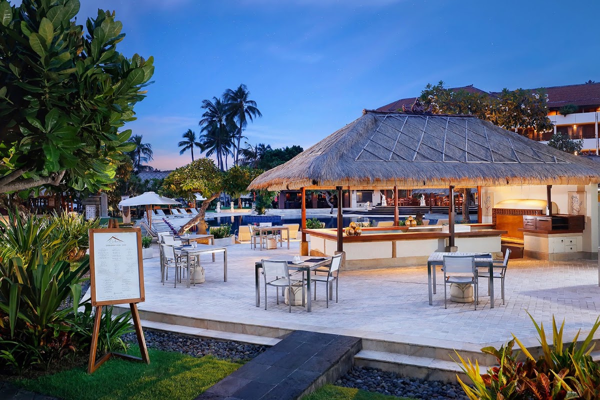 Nusa Dua Beach Hotel & Spa's beachfront dining area at dusk with a thatched-roof restaurant, outdoor tables, pool, and palm trees under a clear blue sky.