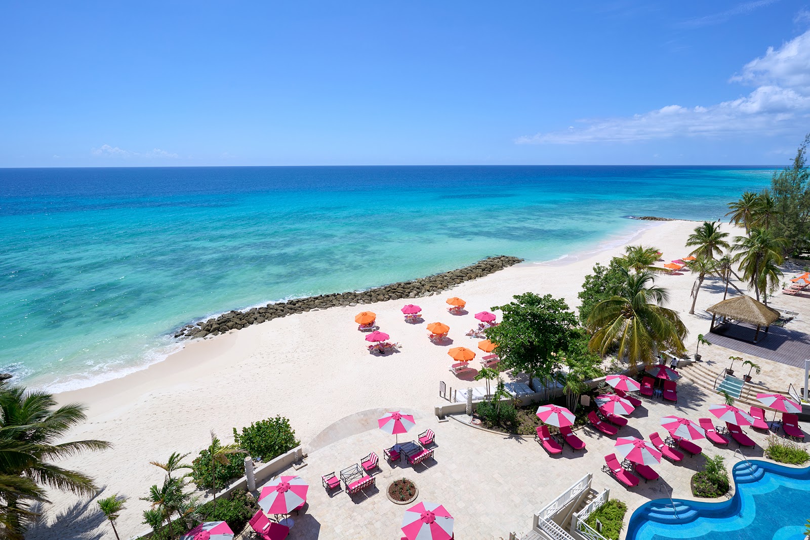 White sand beach with turquoise ocean, pink and orange umbrellas, lounge chairs, palm trees, and a blue pool at O2 Beach Club & Spa.