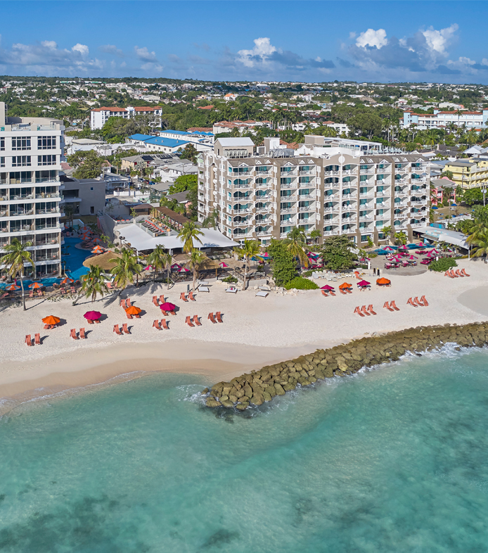 Aerial view of O2 Beach Club & Spa showing sandy beach with colorful loungers, turquoise water, jetty, and multi-story resort buildings with hillside homes beyond.
