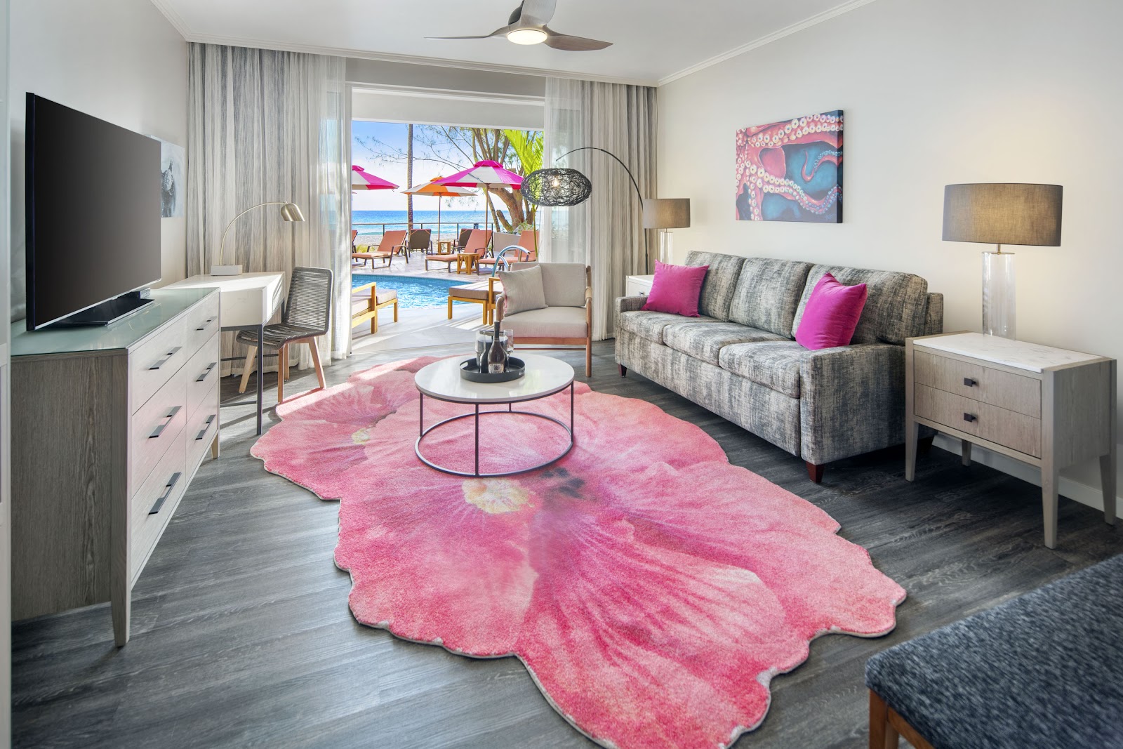 Resort suite living room with gray sofa, pink patterned rug, and ocean view through open doors to pool and beach beyond.