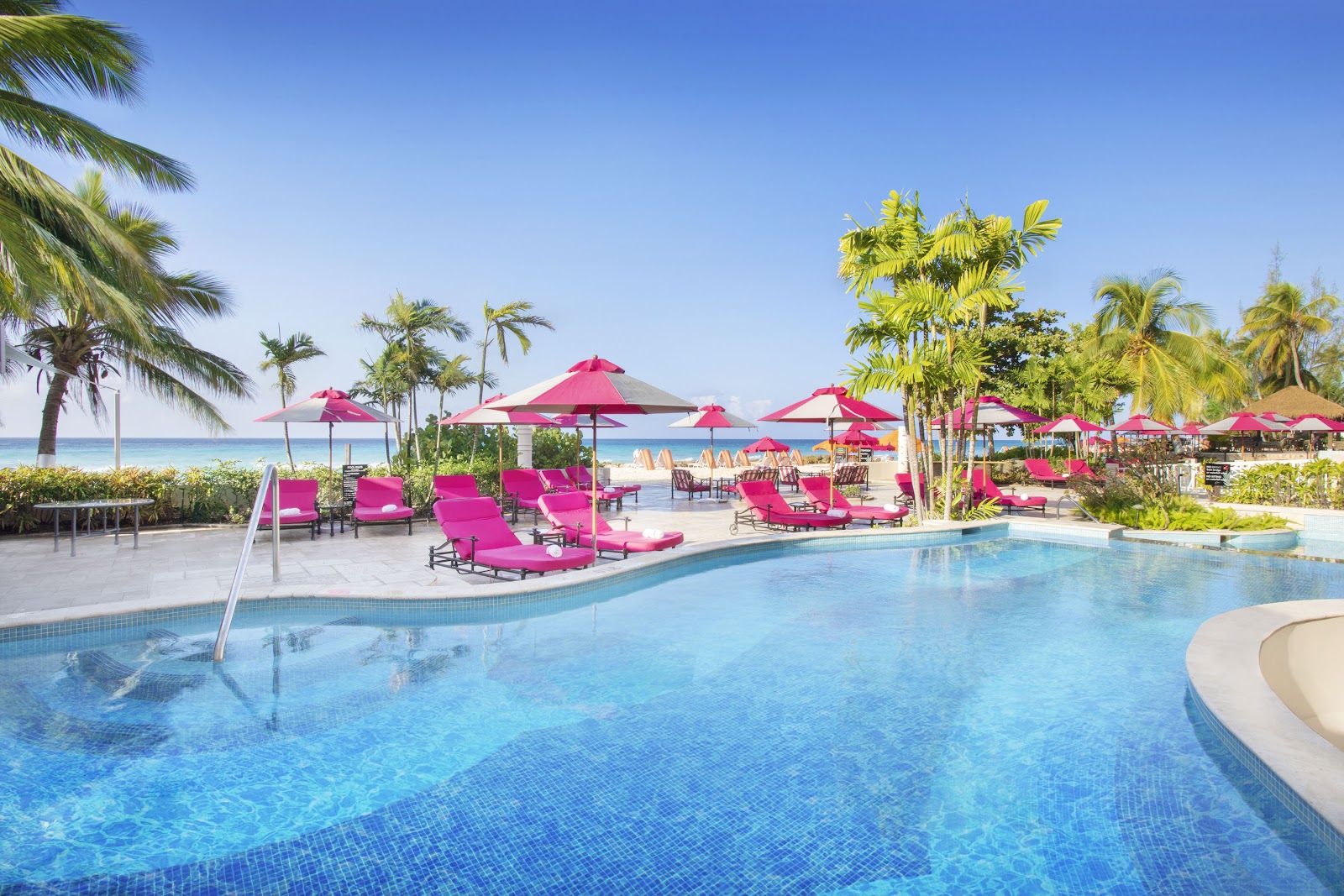 Large resort pool with bright pink loungers and umbrellas, palm trees, and ocean view beyond at O2 Beach Club & Spa.