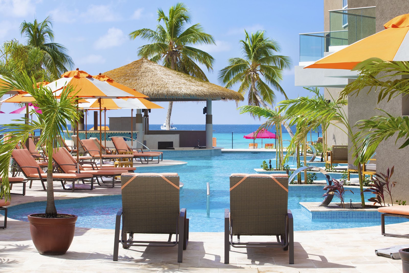 Resort pool with yellow umbrellas, loungers, palm trees, thatched-roof bar, and turquoise ocean beyond under blue sky.