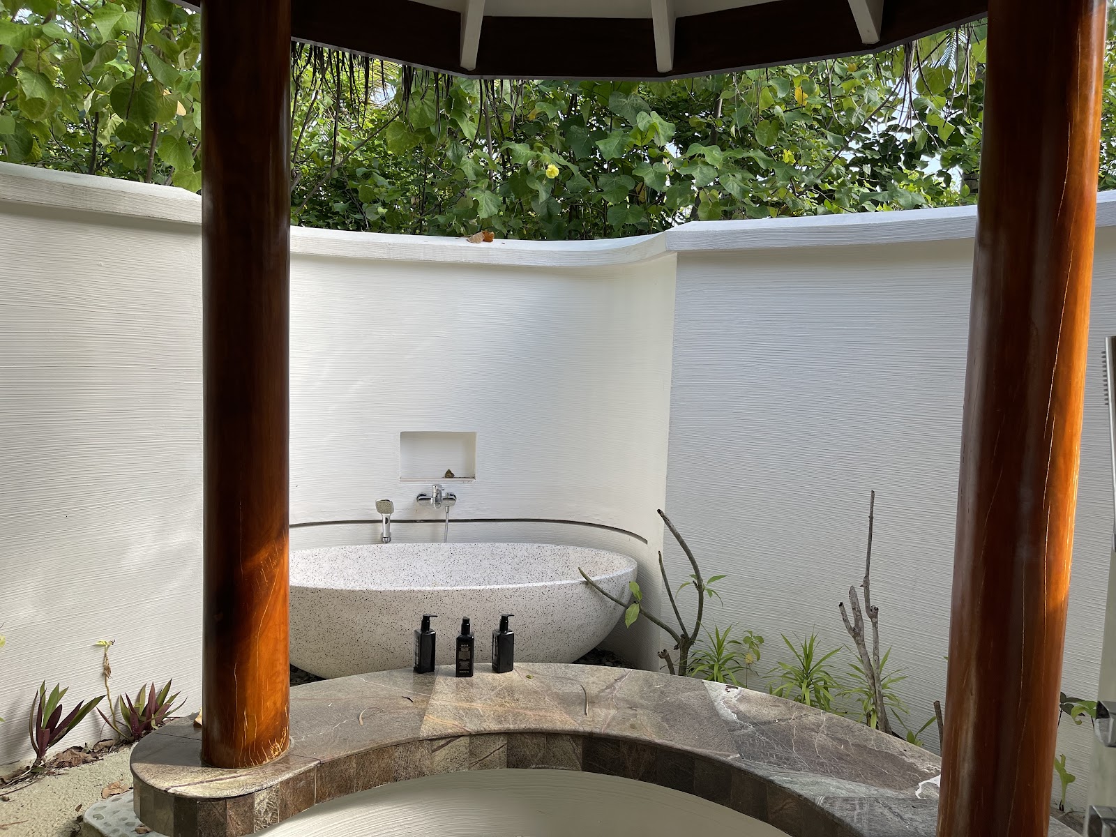 Open-air bathroom with round stone soaking tub surrounded by white walls and wooden posts, tropical plants visible overhead.