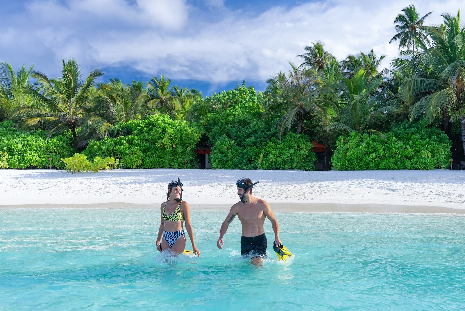 Two guests in snorkeling gear wade into turquoise water at OBLU SELECT Sangeli's white sand beach, backed by lush tropical vegetation and palm trees.