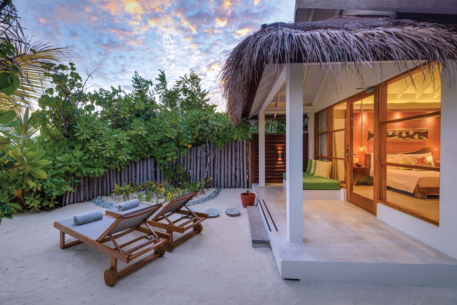 Private beach bungalow at OBLU SELECT Sangeli with thatched roof, wooden loungers on sand, and illuminated interior at dusk.