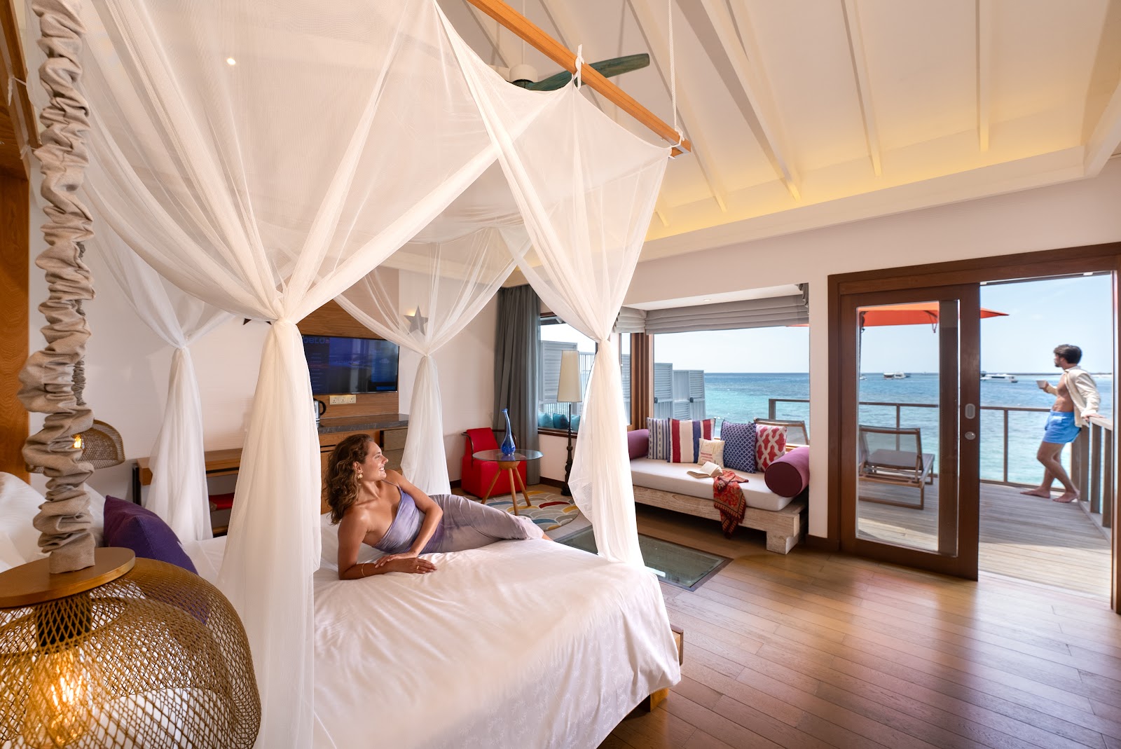 Woman relaxes on a four-poster canopy bed in an ocean-view suite at OBLU SELECT Sangeli, with turquoise water visible through glass doors.