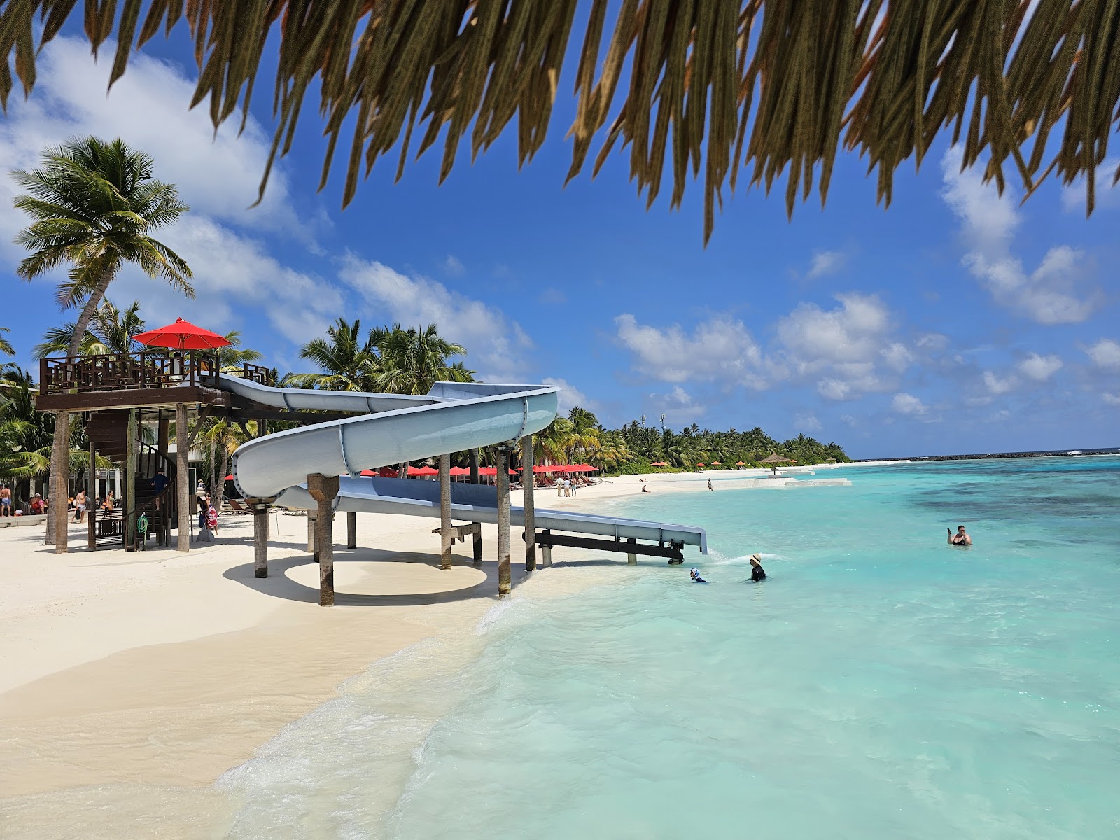 Beach slide and water activities station under thatched roof at OBLU SELECT Sangeli, with turquoise lagoon and palm trees beyond.
