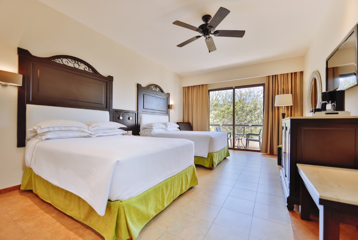 Resort guest room with two beds with white linens and yellow skirts, dark wood headboards, ceiling fan, and balcony doors overlooking trees.