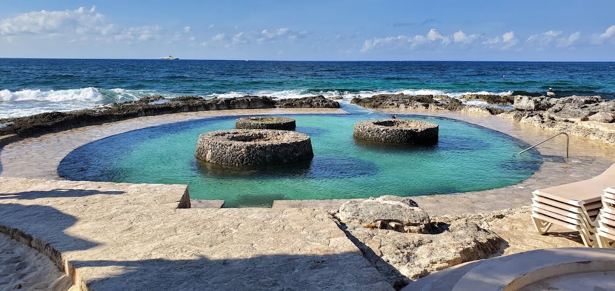Natural rock formations create a teal lagoon pool on a sandy beach at Occidental at Xcaret Destination, with ocean waves beyond.