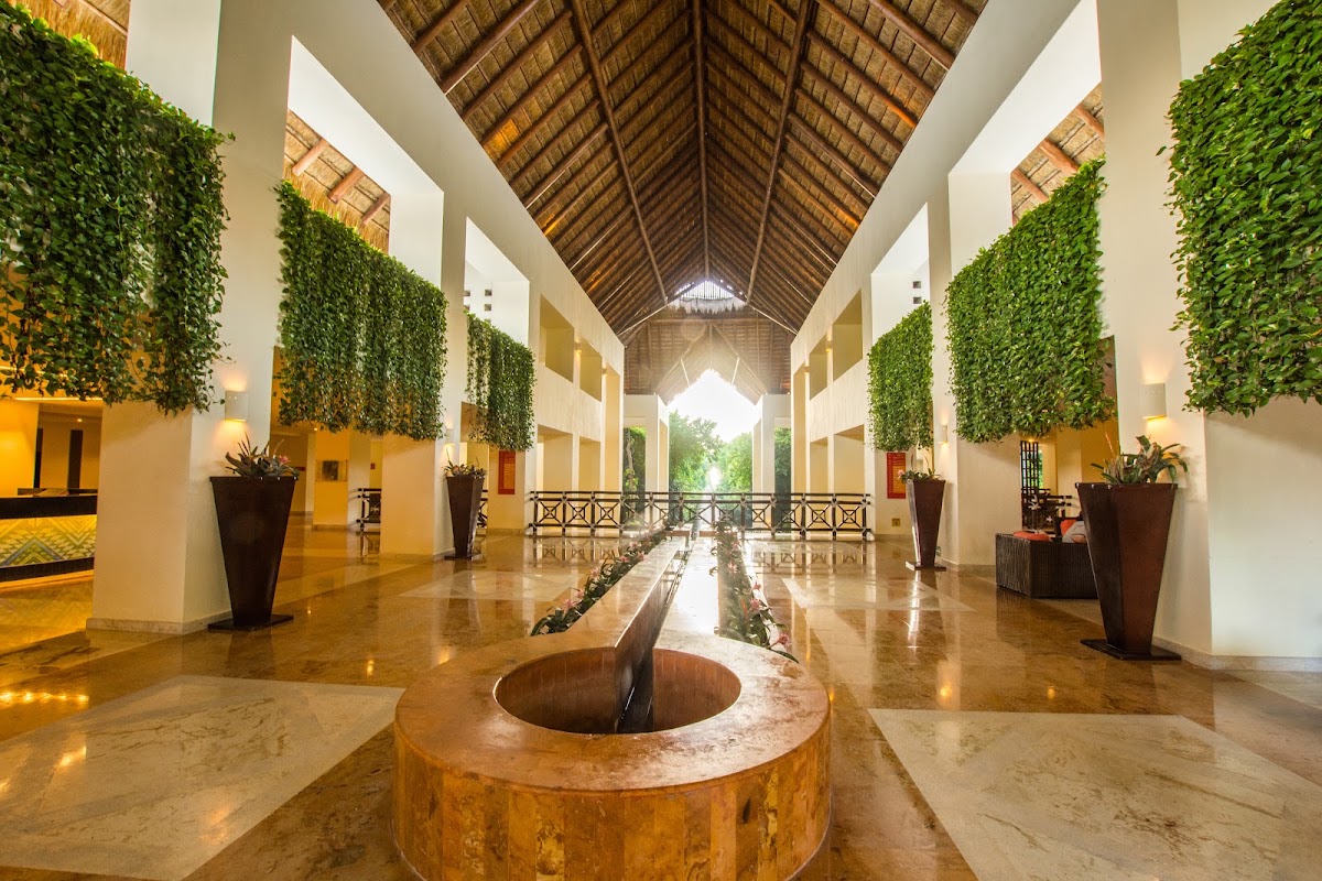 Symmetrical covered corridor with soaring thatch roof, polished stone floor, cascading ivy walls, and central circular fountain opening to ocean view beyond.