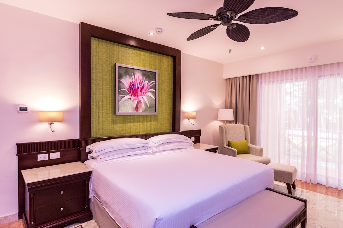 Resort bedroom with queen bed, dark wood headboard with yellow-green fabric panel and pink flower artwork, ceiling fan, bedside lamps, and sliding glass doors with sheer curtains.
