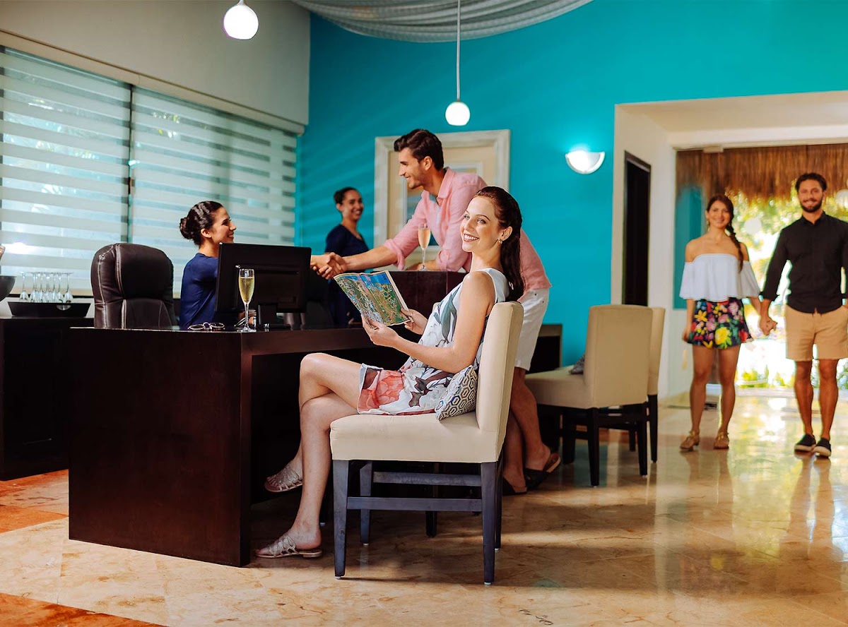 Woman holds resort brochure at turquoise-walled lobby desk while staff and guests move about the modern reception area.
