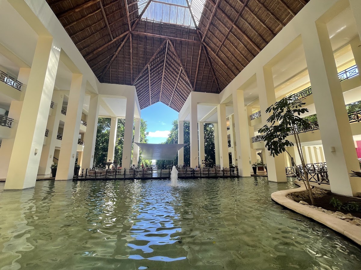 Shallow thermal pool with fountain inside a high-ceilinged atrium at Occidental at Xcaret Destination, cream columns supporting dark wooden roof structure.