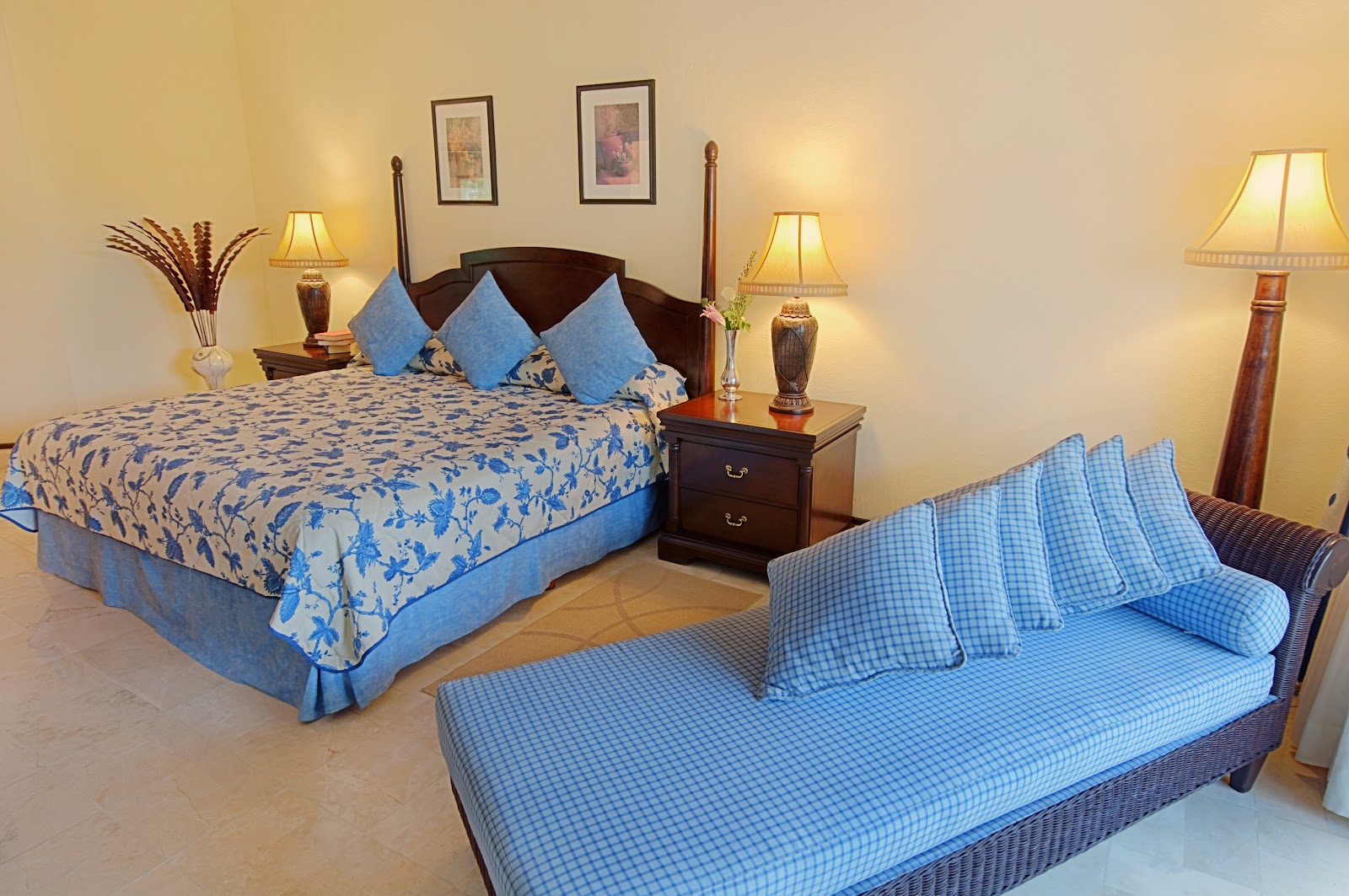 Resort bedroom with blue and white bedding on a four-poster bed and daybed, dark wood nightstand, framed artwork, and warm table lamps on cream walls.