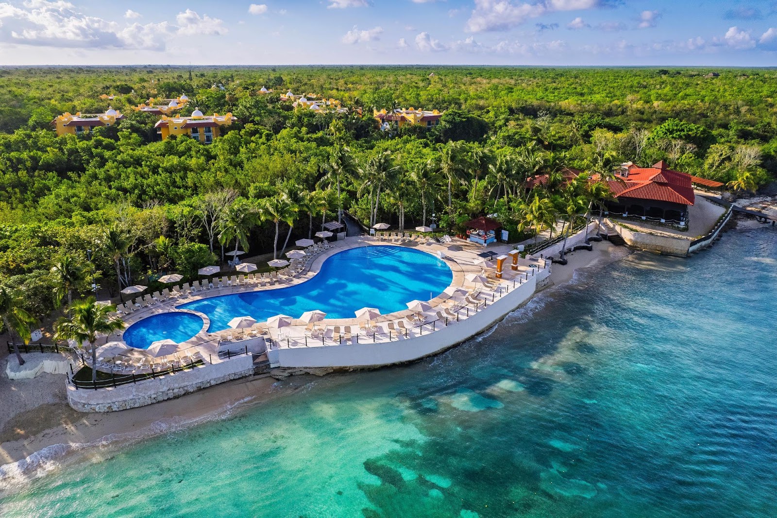 Resort pool with curved shape and lounge chairs sits on beachfront beside turquoise Caribbean water and dense jungle canopy.