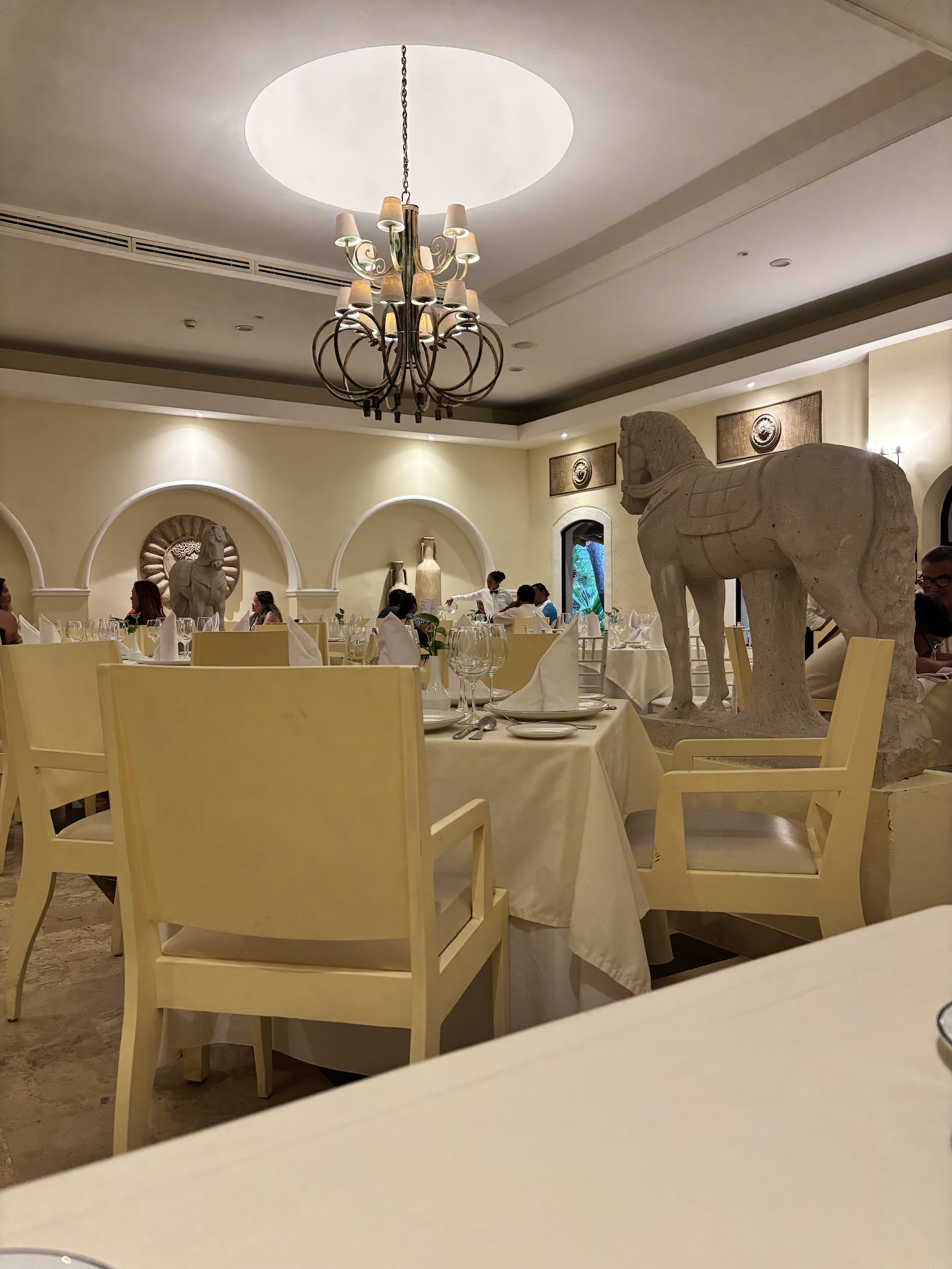 Elegant dining room with cream-colored chairs, white-draped tables, stone elephant sculpture, and chandelier under coffered ceiling.