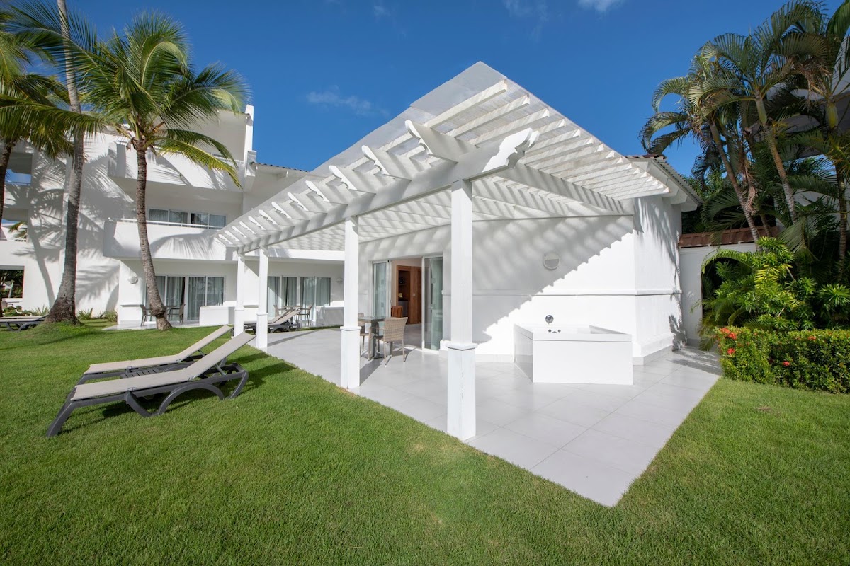 White bungalow with pergola shading a patio, lounge chair on lawn, palm trees and resort buildings visible under blue sky.