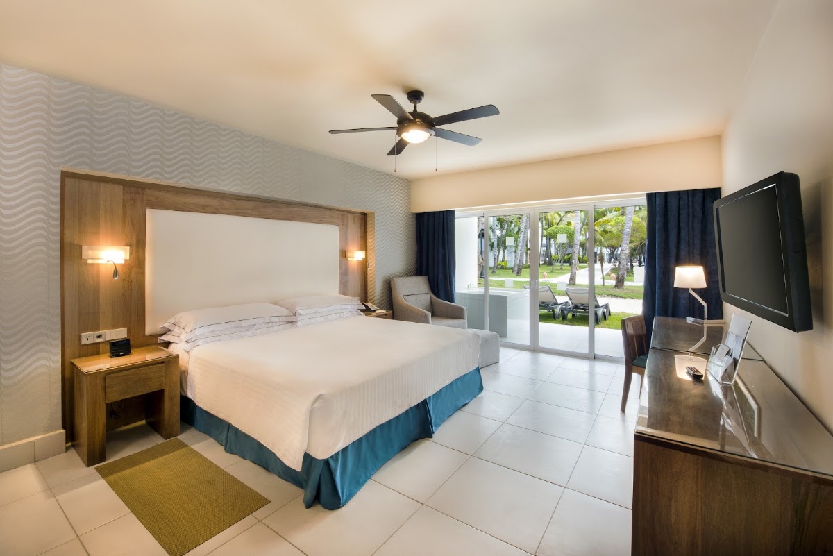 Resort guest room with queen bed, wooden headboard with sconces, flat-screen TV, and glass doors opening to palm-lined grounds.