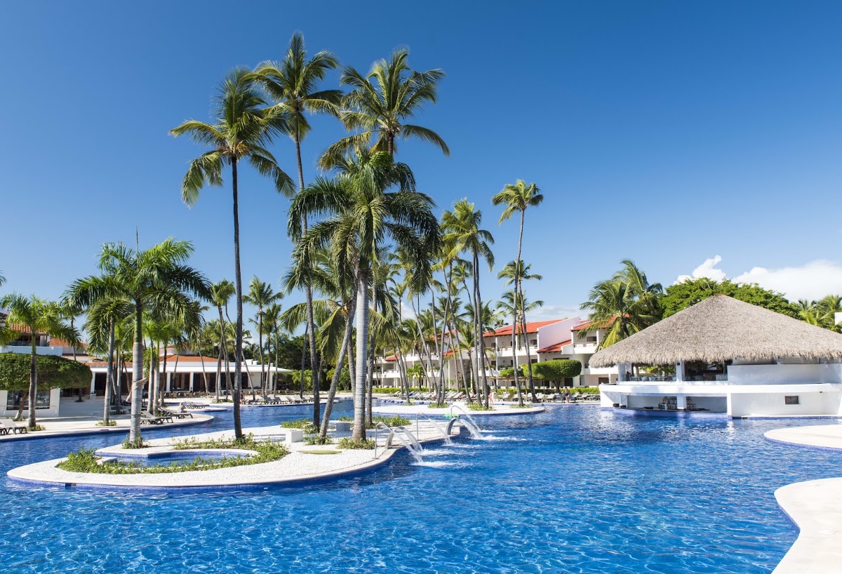 Large resort pool with palm trees, white buildings with terracotta roofs, and a thatched-roof bar structure under clear blue sky.
