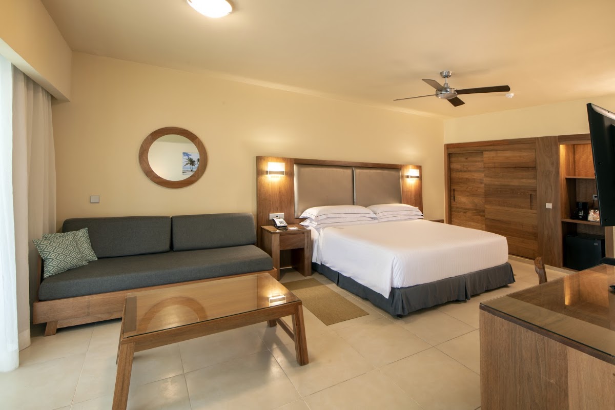 Resort guest room with queen bed, gray sofa, wooden furniture, and beige tile flooring.