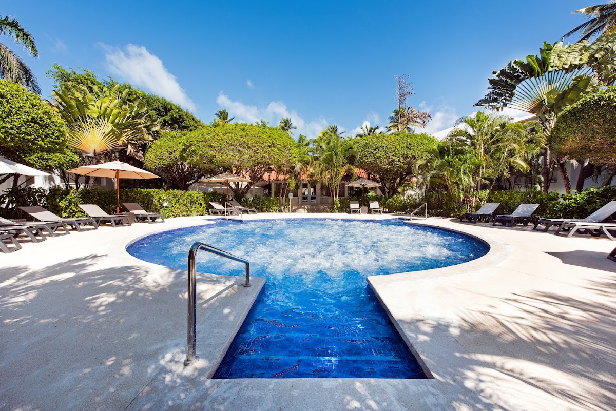 Circular blue pool surrounded by white loungers and tan umbrellas, with lush tropical vegetation and resort buildings in the background.