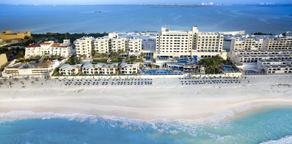 Aerial view of Occidental Tucancún beachfront resort with white sand, turquoise water, multi-story buildings, and blue lounge chairs lining the beach.