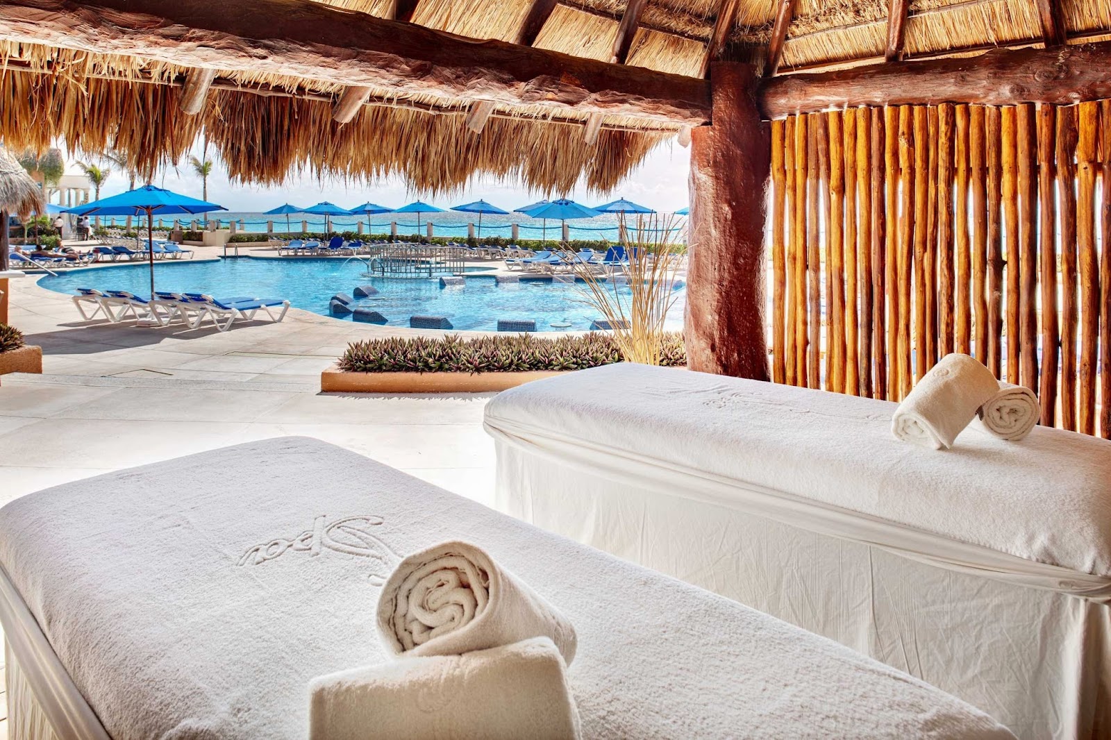 Spa treatment beds with rolled towels overlook a resort pool with blue umbrellas and ocean views beyond.